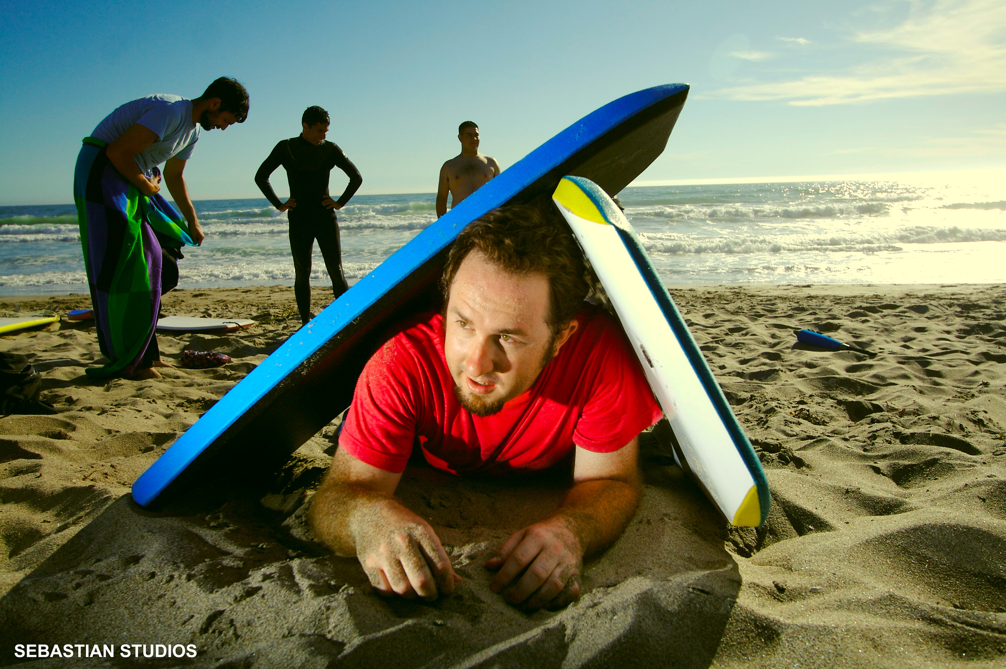 The man lies on the beach. Two surfboards lie above his head. Three men stand in the background